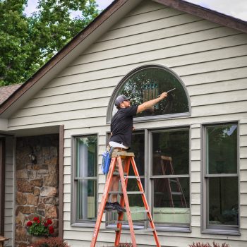 Young man stands on ladder holding squeegee while washing outside windows of Midwestern house in summer